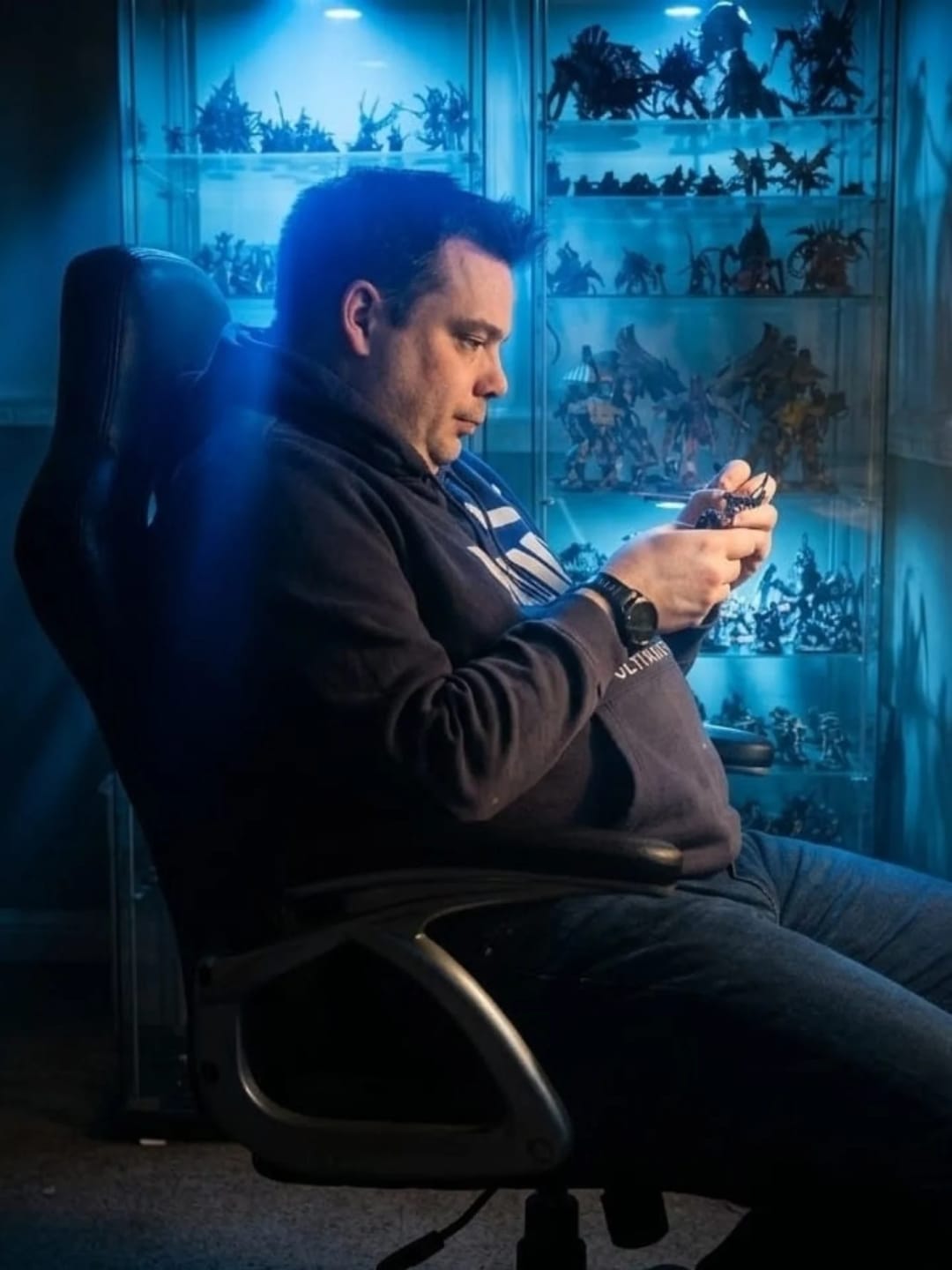 Chris of Brooko's Forge painting a miniature at his bench in front of a lit display cabinet